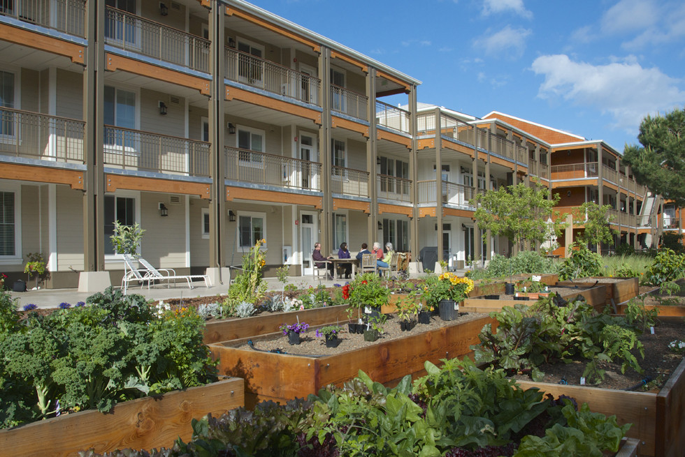 A view of a three-storey housing development with a veggie garden in front of it. Each level is surrounded by a wide veranda. A group of people sits at a table talking in front of the buildings.