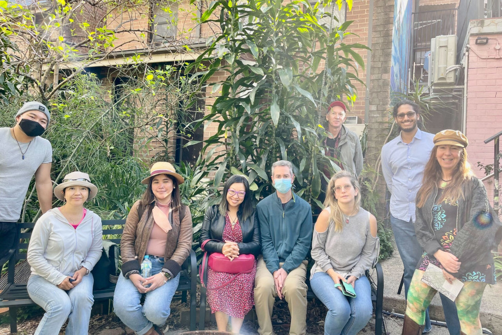 A group of Sydney Cohousing members sitting in the garden at Alpha House. Some are wearing masks, hats, or glasses. It's a sunny day. Behidn the group are tall green leafy plants, and brick walls.